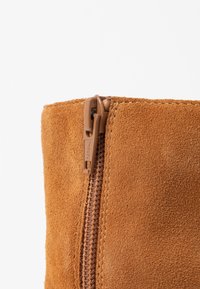 Botín de ante en marrón con una textura suave, que presenta una cremallera marrón y el logotipo de YKK en el tirador de la cremallera.