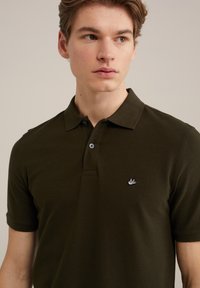 Tamsiai žalia polo marškinėliai, trumpomis rankovėmis, juostele apdailinta apykakle, dviem mygtukais užsegami, su mažo išsiuvinėto logotipo ant krūtinės. Pagaminti iš medvilnės.