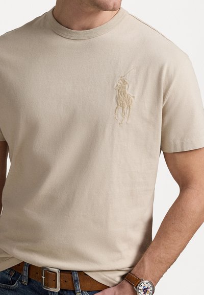 Beige kokvilnas T-krekls ar apaļu kaklu, īsām piedurknēm un teksturētu tonālu izšūtu polo logo kreisajā krūtī.