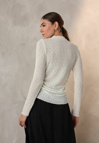 Lightweight cream long-sleeve top with textured pattern and gathered detail at the waist, paired with a black skirt, shown from the back.