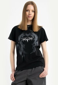 Młoda kobieta z prostymi, jasnobrązowymi włosami, ubrana w czarny t-shirt z grafiką i szare spodnie, stoi z jedną ręką w kieszeni na tle białej ściany.