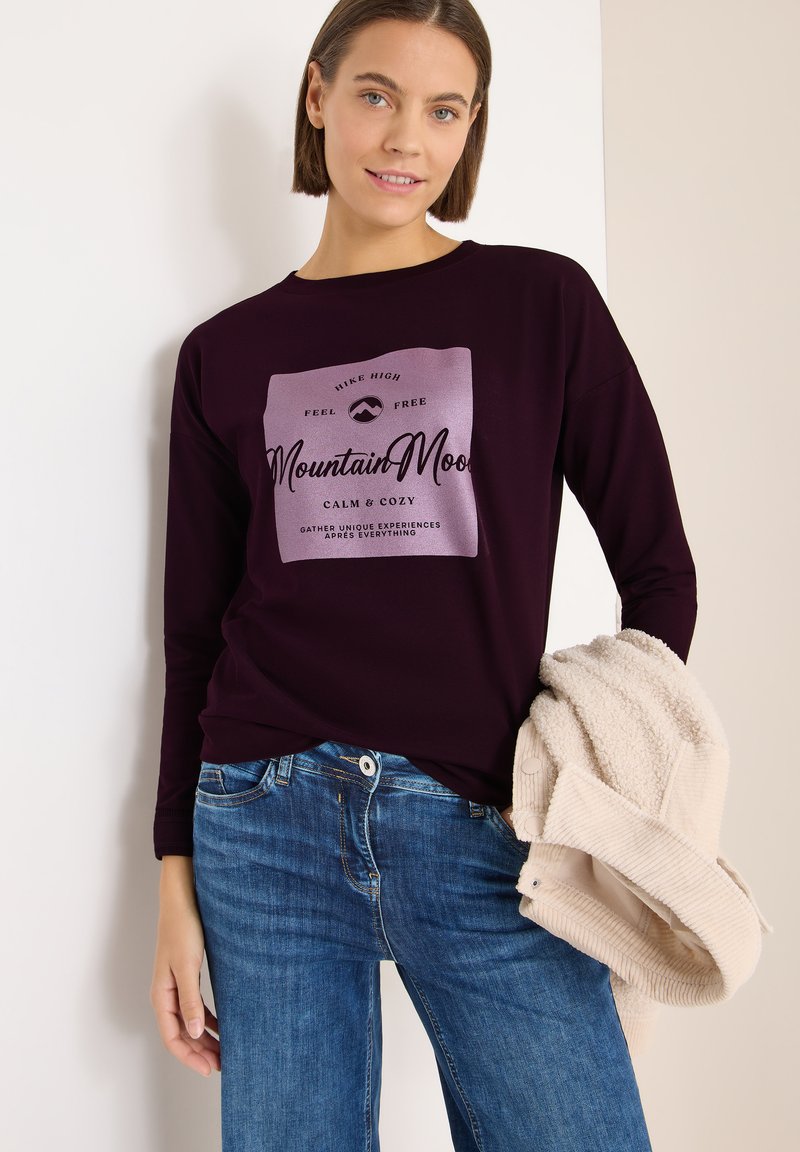 Langärmliges lila T-Shirt mit einem helllila Grafik-Print auf der Vorderseite. Kombiniert mit blauen Jeans und hält eine cremefarbene strukturierte Strickjacke.