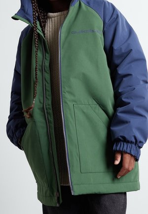 Personne portant une veste zippée verte et bleu marine par-dessus un pull en tricot beige, avec des cheveux tressés tombant sur l'épaule.