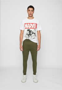 Weißes T-Shirt mit einem großen roten "MARVEL"-Logo und schwarzer Comic-Art, kombiniert mit olivgrünen Jogginghosen und weißen Turnschuhen.