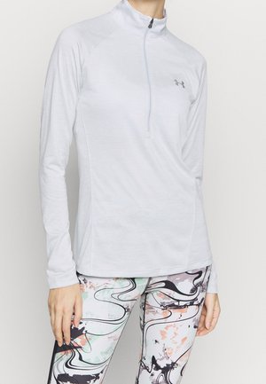 Persoon met een witte sporttop met lange mouwen en ritssluiting in de kraag, en een legging met abstracte zwarte, oranje en groene patronen.
