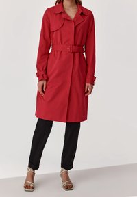 Trench de color rojo con cinturón, con cuello, mangas largas y detalles en los puños, combinado con pantalones negros y sandalias de tiras de color verde claro.