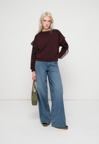 Top burdeos de manga larga con tejido transparente acanalado y volantes, cuello redondo, combinado con vaqueros azul ancho y bolso verde al hombro.