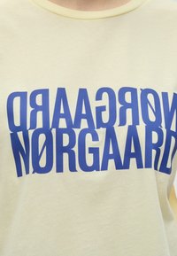 Φωτεινό κίτρινο πουκάμισο με το όνομα της μάρκας "NØRGAARD" τυπωμένο σε μεγάλες, μπλε, παραμορφωμένες κεφαλαία γράμματα στο στήθος.