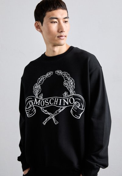 Mann, der einen schwarzen Moschino-Pullover mit weißem Lorbeerkranz und gekreuzten Stöcken trägt, blickt seitlich vor einem schlichten Hintergrund.