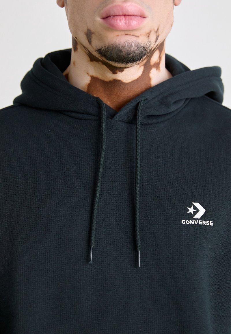 Homme atteint de vitiligo portant un sweat à capuche Converse noir avec un logo sur la poitrine, barbe visible et lèvres rosées claires sur un fond uni.