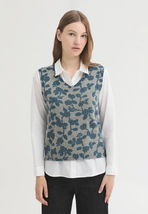 Jeune femme portant une chemise blanche sous un gilet sans manches gris avec un motif floral bleu, debout devant un fond uni.