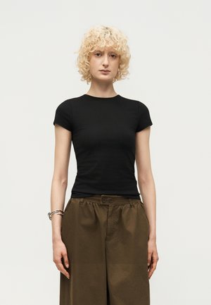Femme aux cheveux blonds courts et bouclés, portant un haut noir ajusté à manches courtes et un pantalon marron ample, debout devant un fond uni.