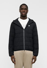 Μαύρο καπιτονέ μπουφάν με φερμουάρ στο μπροστινό μέρος, ρυθμιζόμενο κουκούλα και λογότυπο στο στήθος. Διαθέτει αντίθετες ραφές και ελαστικές μανσέτες.