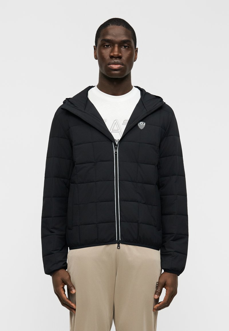 Μ αύρο καπιτονέ μπουφάν με φερμουάρ στο μπροστινό μέρος, ρυθμιζόμενο κουκούλα και λογότυπο στο στήθος. Διαθέτει αντίθετες ραφές και ελαστικές μανσέτες.