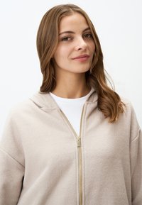 Felpa beige con zip, realizzata in un morbido tessuto con cappuccio, dotata di una zip dorata e di uno strato interno bianco liscio, progettata con una vestibilità comoda.