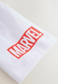 Bílá bavlněná látka s výrazným červeným logem "MARVEL" potisknutým na rukávu. Hladká textura s rovnými švy.