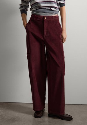 Pantalons en velours côtelé évasés de couleur bordeaux, dotés de stries verticales, d'une fermeture à bouton et de poches latérales. Portés avec des chaussures marron foncé.