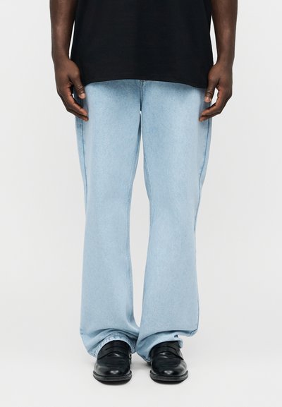 Mężczyzna w jasnobłękitnych szerokich dżinsach, czarnych butach i czarnej koszuli, stojący na tle jednolitej białej ściany.