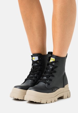 Bottes chevilles en cuir noir à finition texturée, dotées d'une semelle beige épaisse, d'un bout rond et de lacets noirs. Étiquettes jaunes remarquables sur la languette.