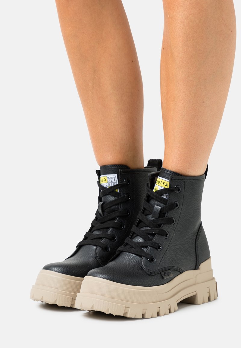 Bottes chevilles en cuir noir à finition texturée, dotées d'une semelle beige épaisse, d'un bout rond et de lacets noirs. Étiquettes jaunes remarquables sur la languette.