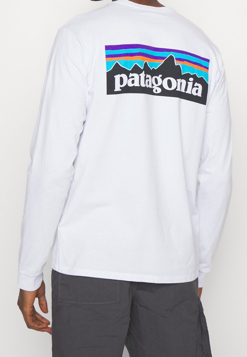 Valkoinen pitkähihainen t-paita, jossa on värikäs vuori graafinen kuva ja "patagonia"-teksti selässä. Puuvillakangasta, ribatut hihanpäät ja suora helma.