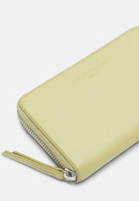 Żółty skórzany portfel o gładkiej fakturze, wyposażony w zamek błyskawiczny i wytłoczoną nazwę marki na przodzie. Zawiera dopasowany materiałowy uchwyt zamka.