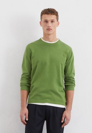 Pull en maille verte avec un col rond et des manches longues, doté de poignets et d'un ourlet côtelés. Porté par-dessus un t-shirt blanc à col rond.