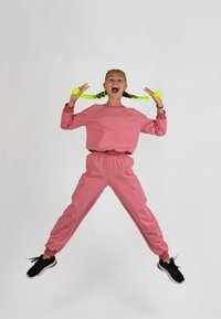 Rosa Sweatshirt und passende Hose mit Seitentaschen. Modell trägt schwarze Sneaker. Haare in Zöpfen mit neon-grünen Akzenten. Sprungpose.
