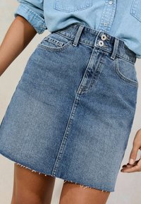 Jeans krilo s čupavim robom, svetlo modre pranje, sprednja gumbna zanka na sredini in stranske žepe. Material ima subtilno teksturo in šivanje.