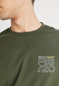 Olīvu zaļš kokvilnas T-krekls ar apaļu kaklu un īsām piedurknēm, ar baltu un dzeltenu grafiku "RED BULL® RACING RB/20".