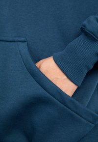 Mano parcialmente insertada en el bolsillo frontal de una sudadera azul marino con puño acanalado visible.