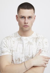 Camiseta blanca de manga corta con un estampado tropical en beige que incluye palmeras y paisajes. Modelo llevando una pulsera de cadena plateada.