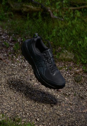 Chaussure de sport noire suspendue au-dessus d'un sentier en gravier avec de l'herbe verte et du feuillage en arrière-plan.