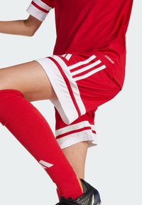 Maillot de sport rouge avec des rayures blanches, associé à un short rouge avec des accents blancs. Des chaussettes rouges jusqu'aux genoux et des chaussures noires complètent la tenue.