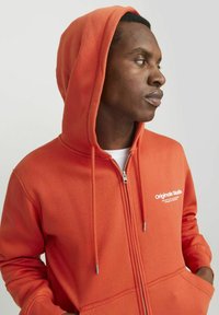 Sudadera con cremallera naranja, con un bolsillo frontal, texto de logo blanco en el pecho y capucha ajustable con cordón. Material suave de mezcla de algodón.