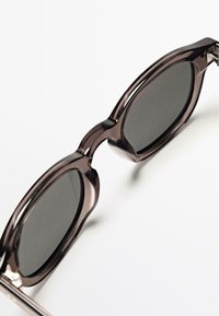 Prohnuté hnědé sluneční brýle s tmavými skly, zaobleným tvarem, silnými obroučkami a kovovými zdobeními na stranách.