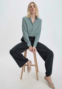 Blusa azzurra chiara con chiusura a bottoni, abbinata a pantaloni a gamba larga neri e ballerine beige, seduta su uno sgabello di legno. Design semplice.