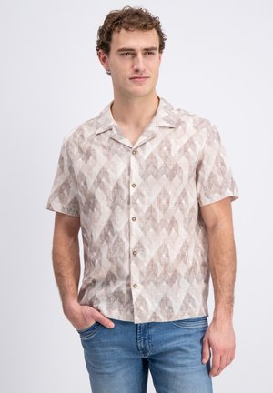 Man met een beige, kortermig overhemd met patroon en een spijkerbroek, staande met één hand in zijn broekzak tegen een lichte achtergrond.