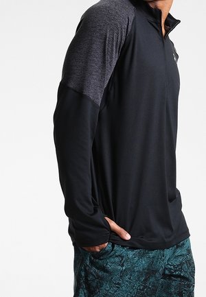 Mann trägt ein schwarzes, langärmliges Sportshirt mit dunkelgrauen Schultern und gemusterten dunkelgrünen Shorts, steht mit einer Hand in der Tasche.