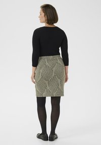 Schwarzes Langarmoberteil, knielanger grüner Rock mit schwarzem wellenförmigem Muster, schwarze Strumpfhose und schwarze Schuhe. Einfaches und figurnahes Design.