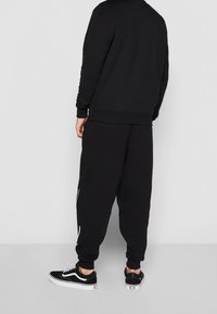 Sudadera negra y pantalones joggers a juego con puños elásticos, que presentan una franja blanca como acento. Zapatillas negras con suela y cordones blancos.