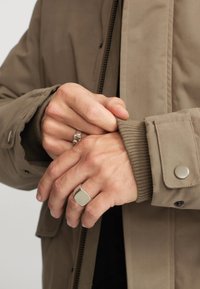 Brun lynlåset jakke med ribbede manchetter, prydet med sølvfarvede knapdetaljer. Hænderne er foldet, hvilket fremhæver teksturerede sølvringe.