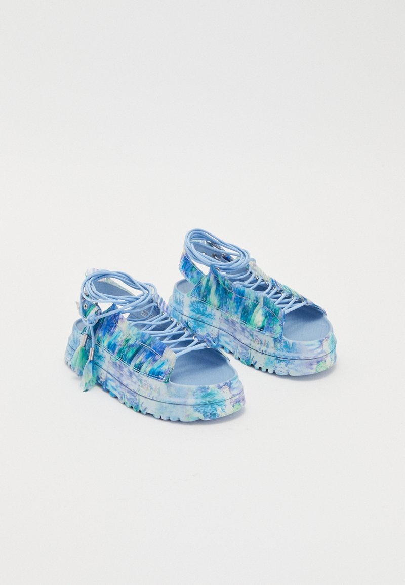 Sandales compensées bleues avec une tige brillante et texturée présentant un motif aquarelle en bleu et vert. Comprend des lacets et des pompons décoratifs.
