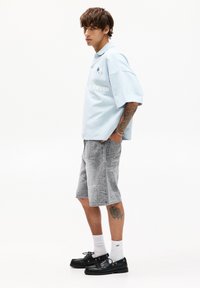 Polo shirt oversized di colore azzurro chiaro con testo stampato bianco, abbinato a pantaloncini di jeans grigi a fantasia. Scarpe nere con lacci e calze bianche.
