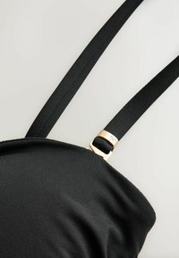 Vêtement en tissu noir avec des bretelles noires réglables et une petite boucle dorée sur un fond clair.