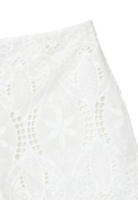 Un tissu blanc brodé présente des motifs floraux et de feuilles avec des détails découpés, transmettant un design texturé et complexe.