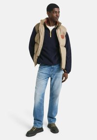 Beige Steppweste über einem marineblauen Pullover, kombiniert mit einer hellblauen Jeans und dunklen Schuhen. Die Accessoires umfassen einen gelben Reißverschluss und ein Patch-Logo.