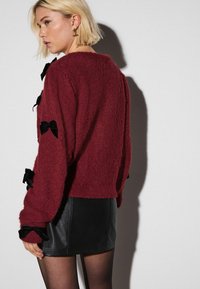 Blond naine, kellel on bob-lõikus, kannab burbooni sviitrit mustade lipsudega ja musta nahast miniseelikut, seistes valge seina ees.