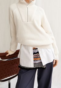 Pull à col montant à fermeture éclair sur le devant, tricoté en côtes couleur crème, superposé sur une chemise blanche et une jupe à motifs, accompagné d'un pantalon sombre.
