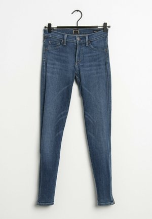 Vaqueros pitillo azules con bolsillos delanteros y trabillas para cinturón colgados de una percha negra sobre un fondo blanco.